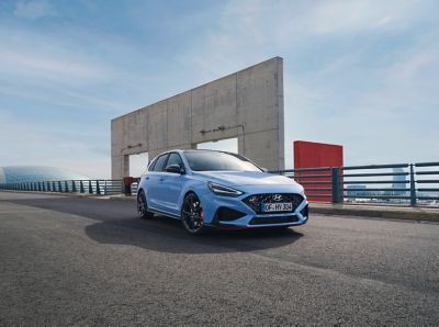 The new Hyundai i30 N from the front right in Performance Blue parked next to an industrial structure.